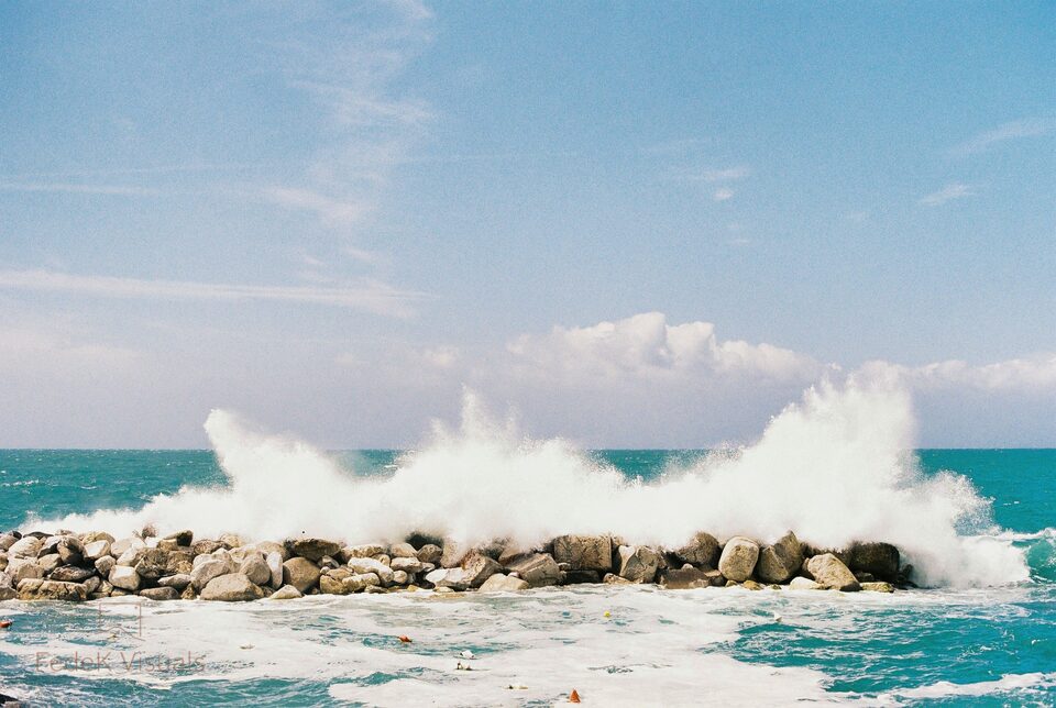 A wave breaking white over a line of sea defence boulders under a soft blue sky.