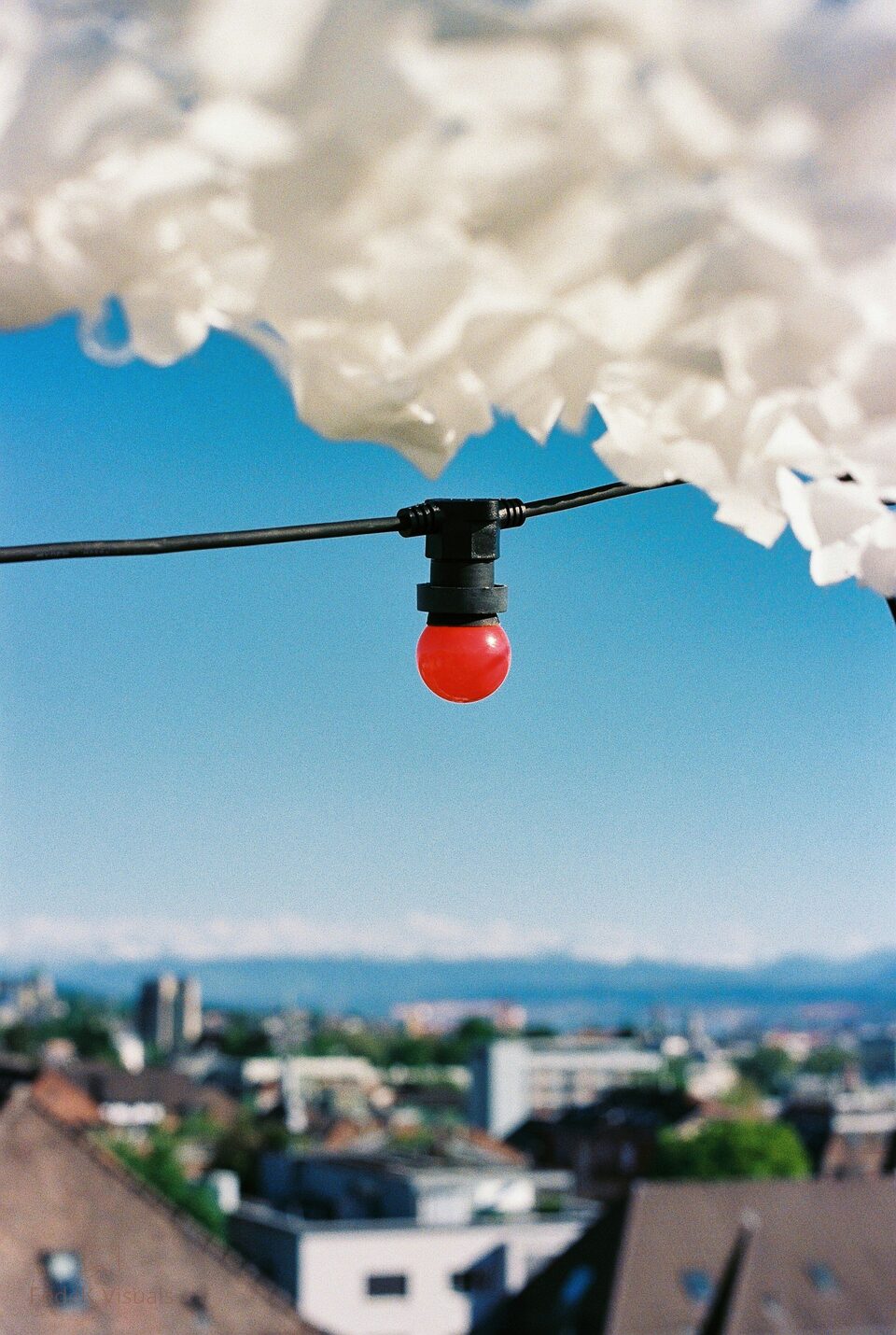 A single red festoon bulb hanging against white cloud, with a town and lake beyond below.