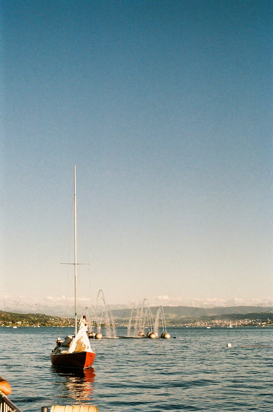 A small sailboat on flat blue water with a hazy far shore and mountains beyond.