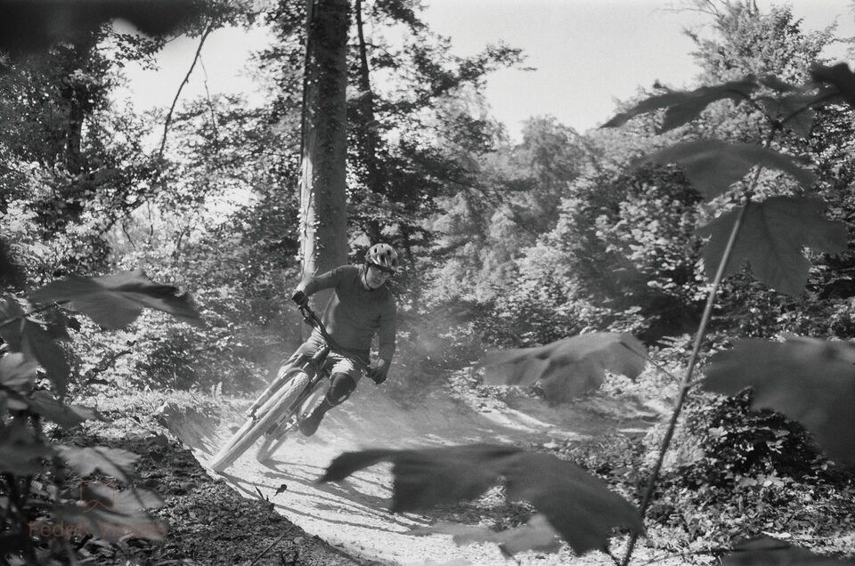 Black and white: a mountain biker railing a bermed forest turn, dust rising, blurred leaves across the foreground.