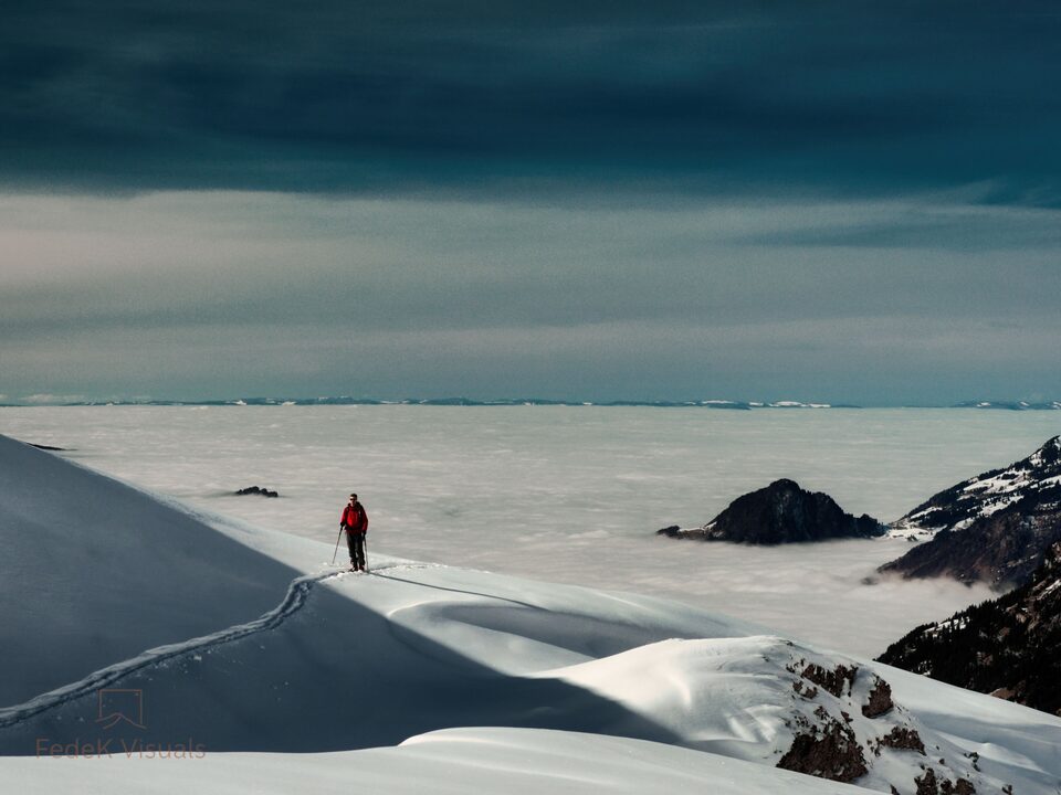 A single figure walking a snow crest above an unbroken sea of cloud, with distant peaks breaking through.