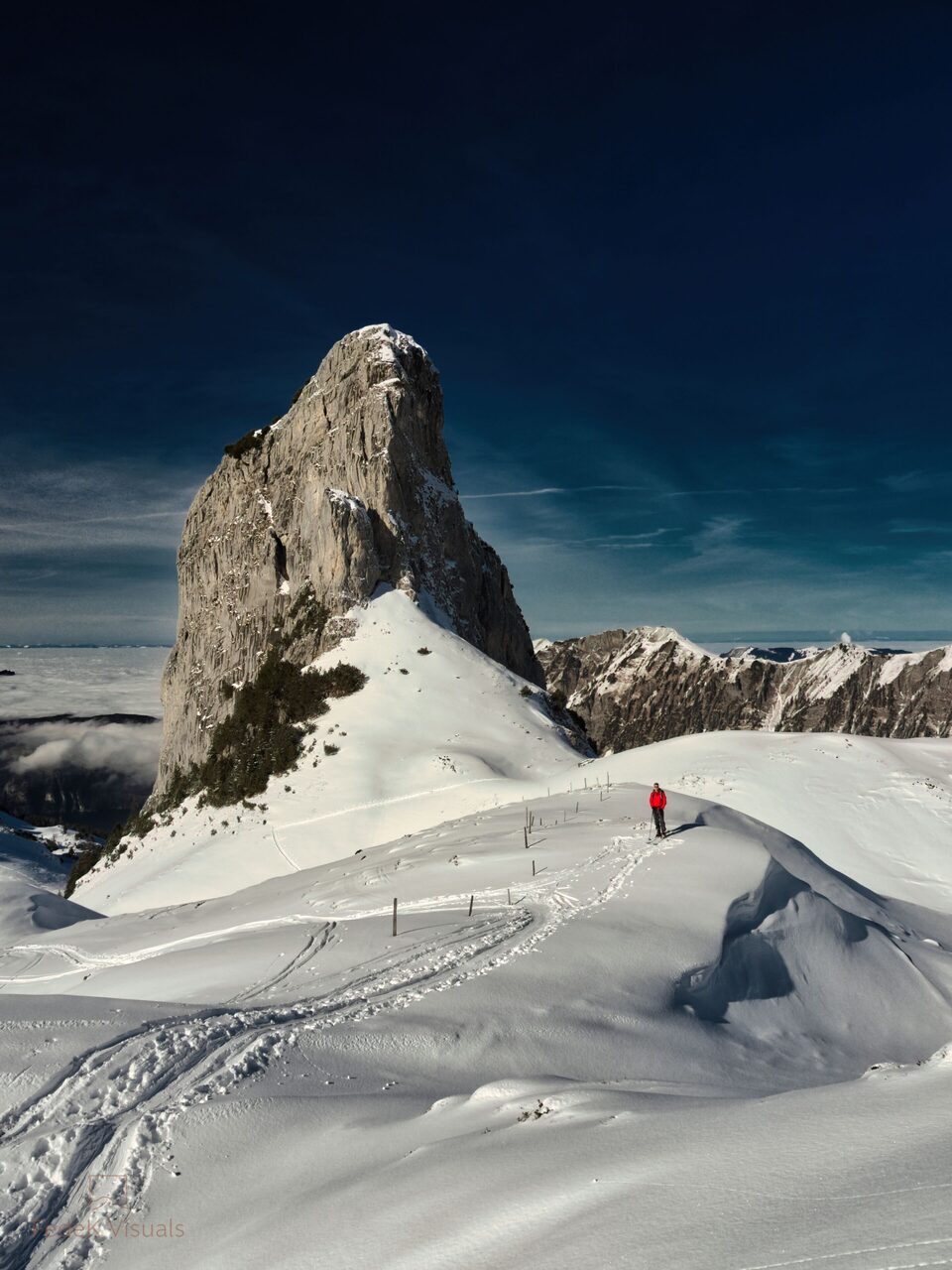 A lone climber in red on a corniced snow ridge, dwarfed by a dark rock summit against deep blue sky.
