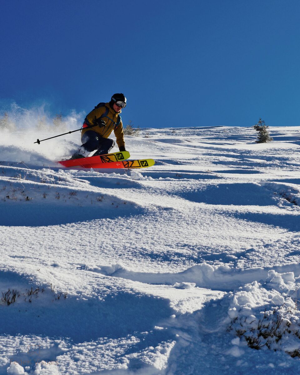 A skier carving through deep powder on a steep open slope below rocky peaks.