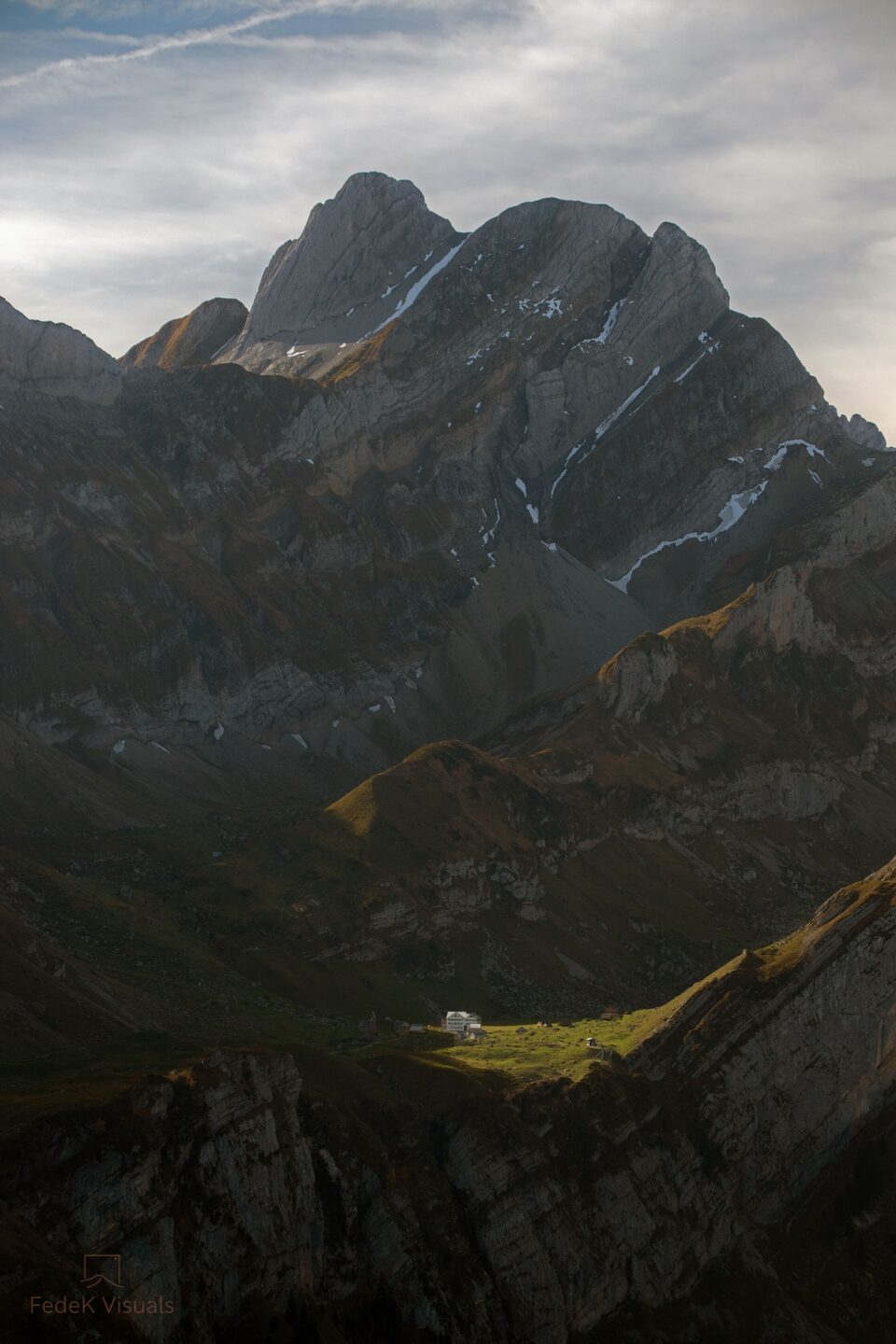 Low light falling across a mountain hut on a broad meadow, with a rugged massif rising behind.
