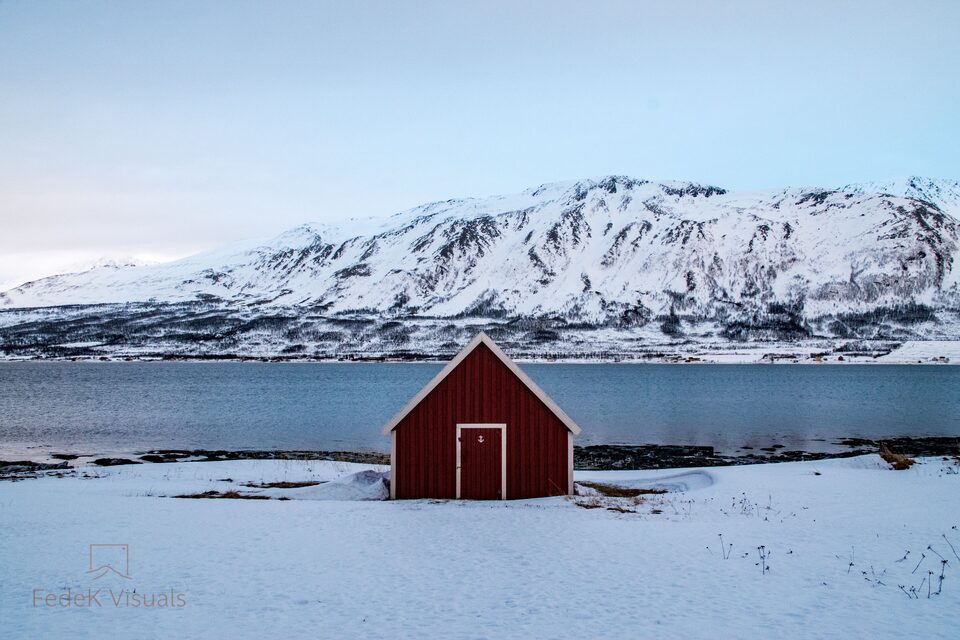A small red wooden hut alone on a snow-covered shore, with a white mountain rising behind it.