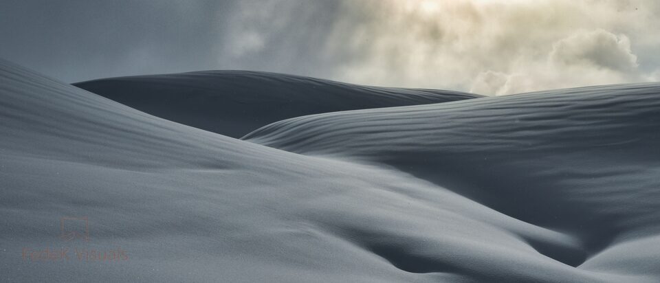 Wind-sculpted dunes of untouched snow in cool grey and blue, with soft cloud breaking behind them.
