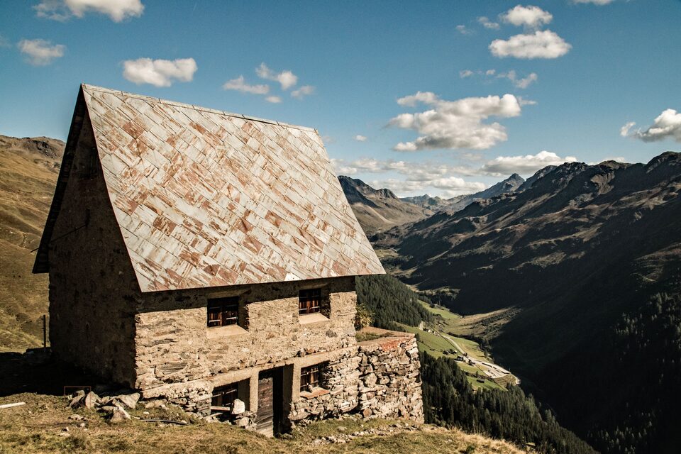 An old stone house with a shingled roof standing on a high ridge above a deep alpine valley.