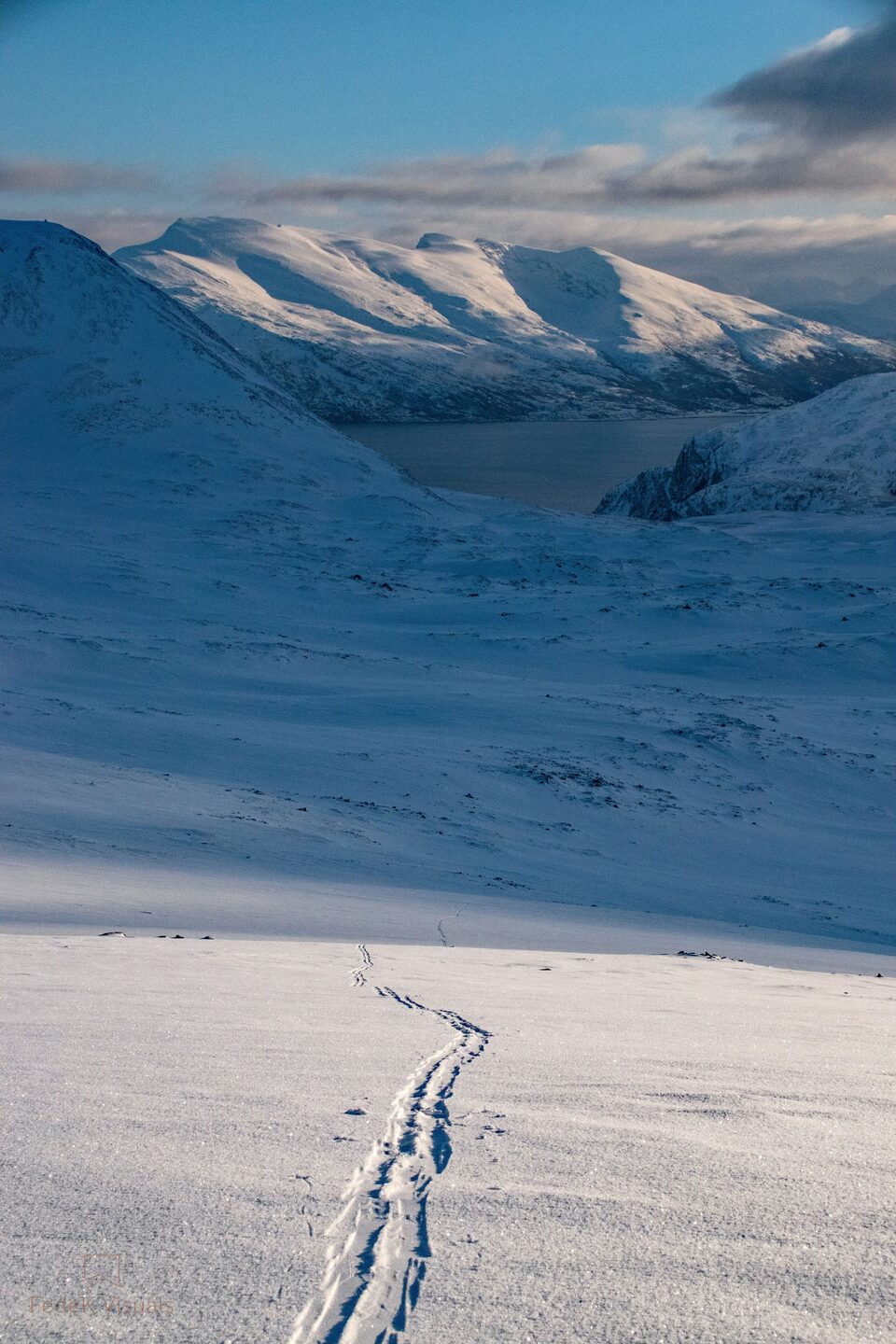 A line of bootprints crossing an empty snowfield towards a long ridge of white mountains.