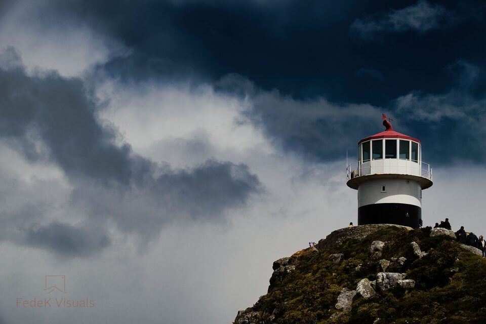 A lighthouse on a rocky headland with heavy cloud rolling in from the left and tiny figures on the rocks below.