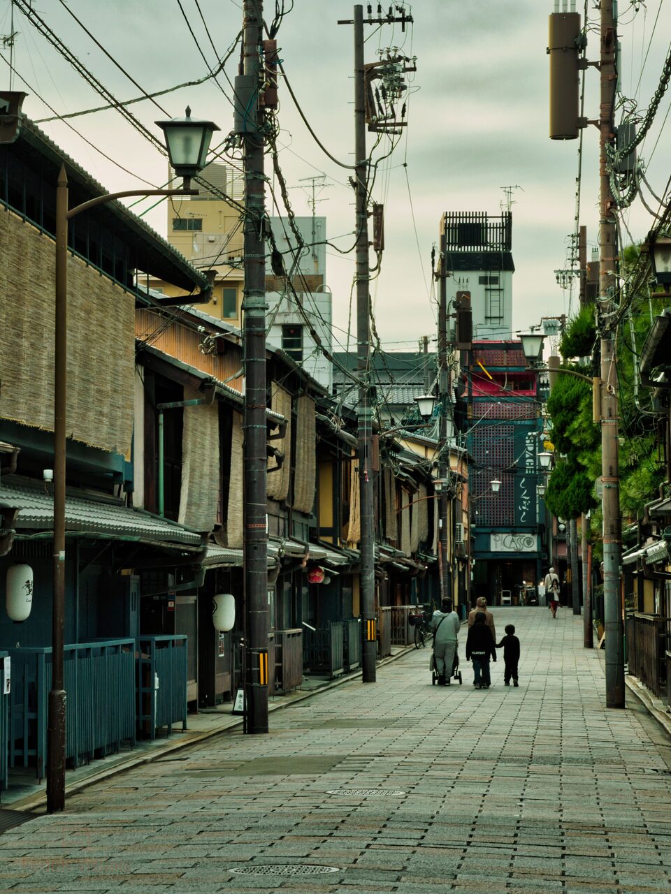 A near-empty Kyoto street of dark wooden machiya houses, two figures small at the far end.