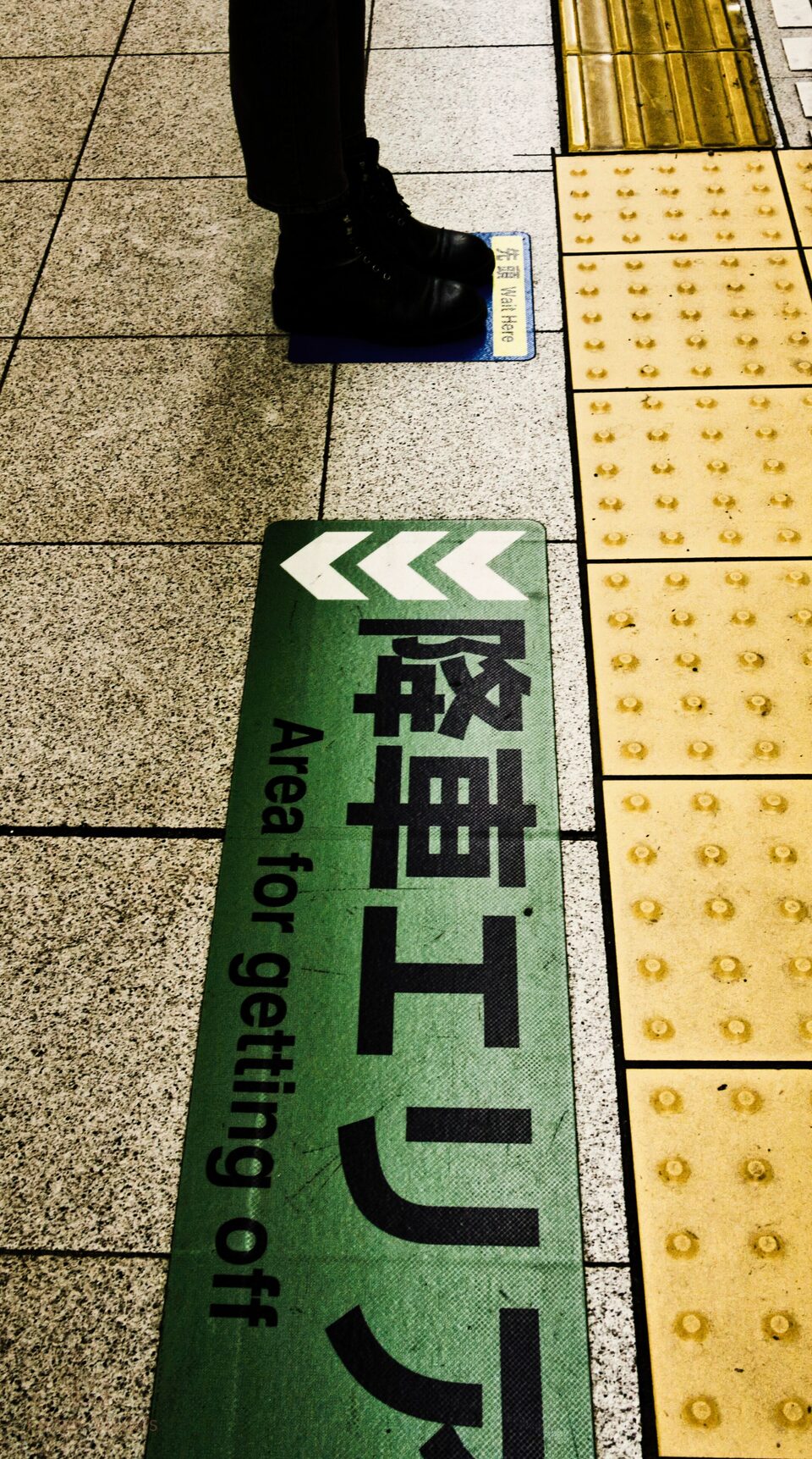 A boot stepping onto a station platform beside a green directional arrow and yellow tactile paving.