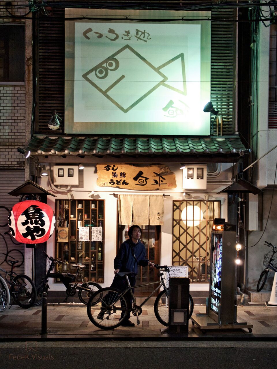 A lit shopfront at night with a red paper lantern and a figure pausing outside.