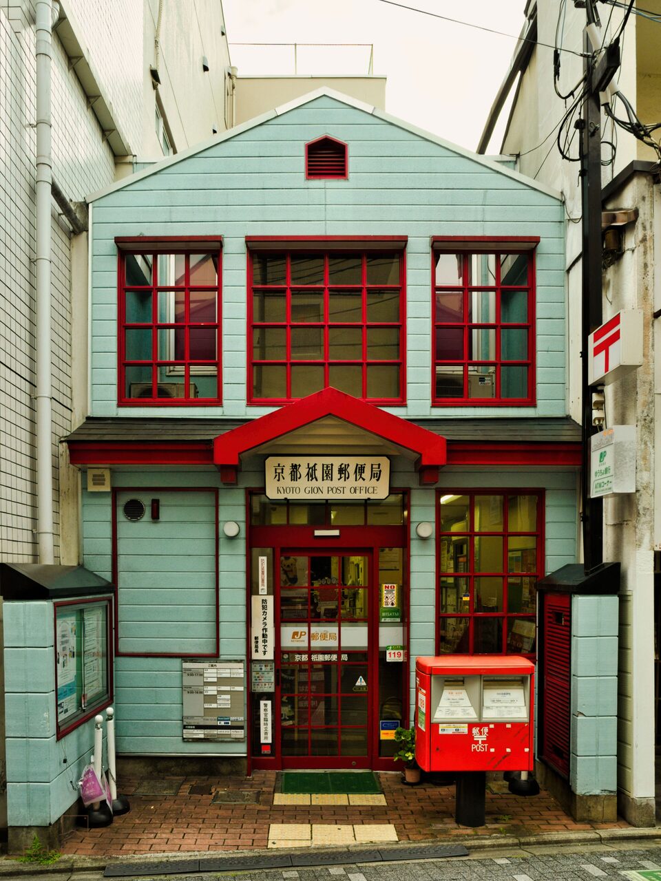 A mint-green wooden post office with red window frames and a red postbox on the pavement.