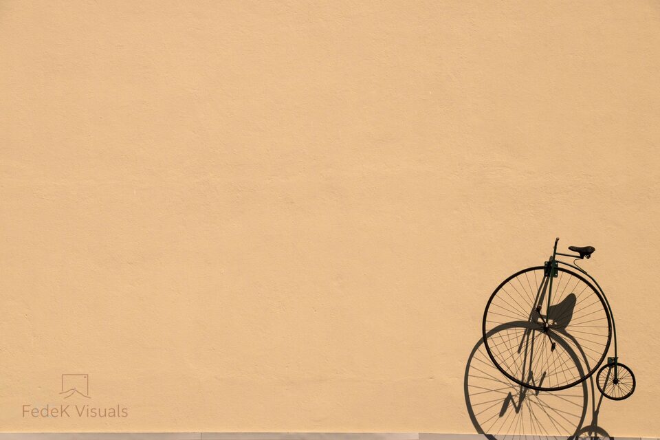 A penny-farthing bicycle standing against an enormous plain beige wall that fills the frame.