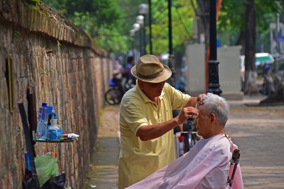 A street barber cutting an older man's hair against a bamboo fence on a city pavement.